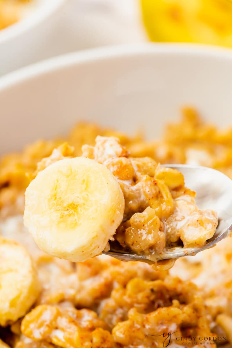 white bowl with golden brown oats with some white milk. Yellow sliced bananas on top then on a spoon a close up of one banana slice and oats