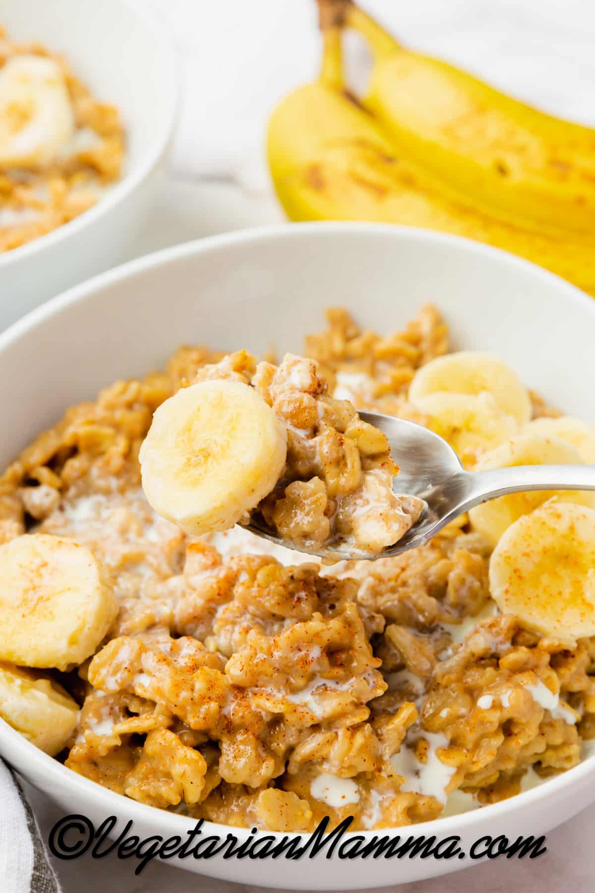 white bowl with golden brown oats with some white milk. Yellow sliced bananas on top then on a spoon a close up of one banana slice and oats