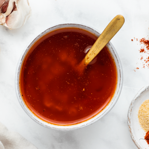 red color liquid in a white bowl with a gold spoon in it. White marble background with yellow and red spices