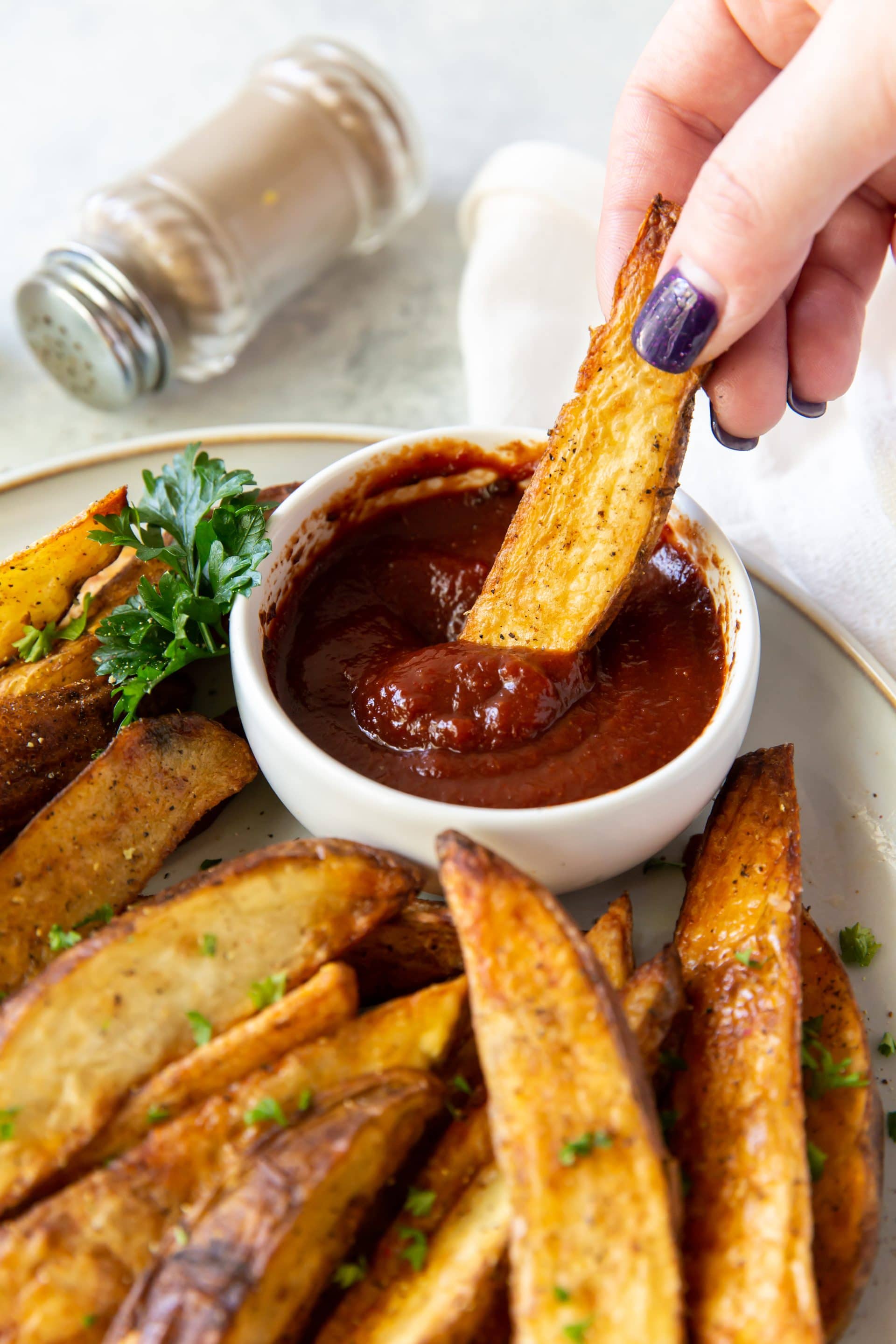 a plate of seasoned, cooked potato wedges with a side cup of ketchup., hand with purple nail dipping fry into ketchup