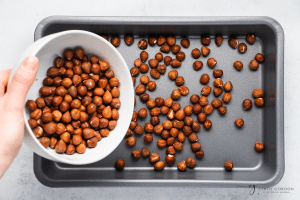 raw hazelnuts being poured from a white bowl into a grey baking pan