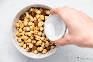 white bowl with roasted hazelnuts and hand pouring salt out of a smaller white bowl