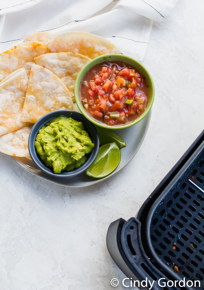 Air Fryer Quesadilla on white plate with guac in a blue bowl and red salsa in a green bowl and a black air fryer basket barely showing to the right
