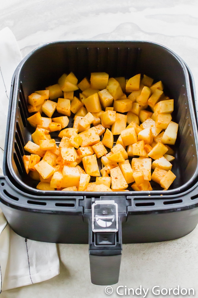 black air fryer basket with raw cubed potatoes with oil and red seasoning on the cubes