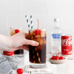 vodka and coke in a tall clear glass with ice and cherries and two straws. Coke can and vodka bottle in back ground. Along with a second vodka coke and a cherry on the table.