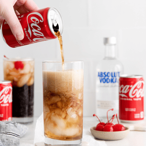 vodka and coke in a tall clear glass with ice and cherries and two straws. Coke can and vodka bottle in back ground. Along with a second vodka coke and a cherry on the table.