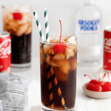 vodka and coke in a tall clear glass with ice and cherries and two straws. Coke can and vodka bottle in back ground. Along with a second vodka coke and a cherry on the table.