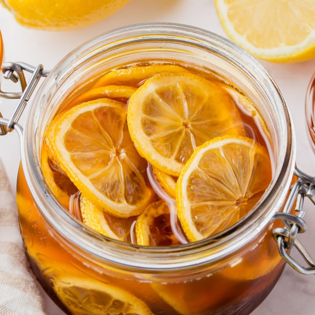 clear jar with lid. Inside are sliced lemons in brown dark honey. slices halved lemons on side and above and honey jar also visable