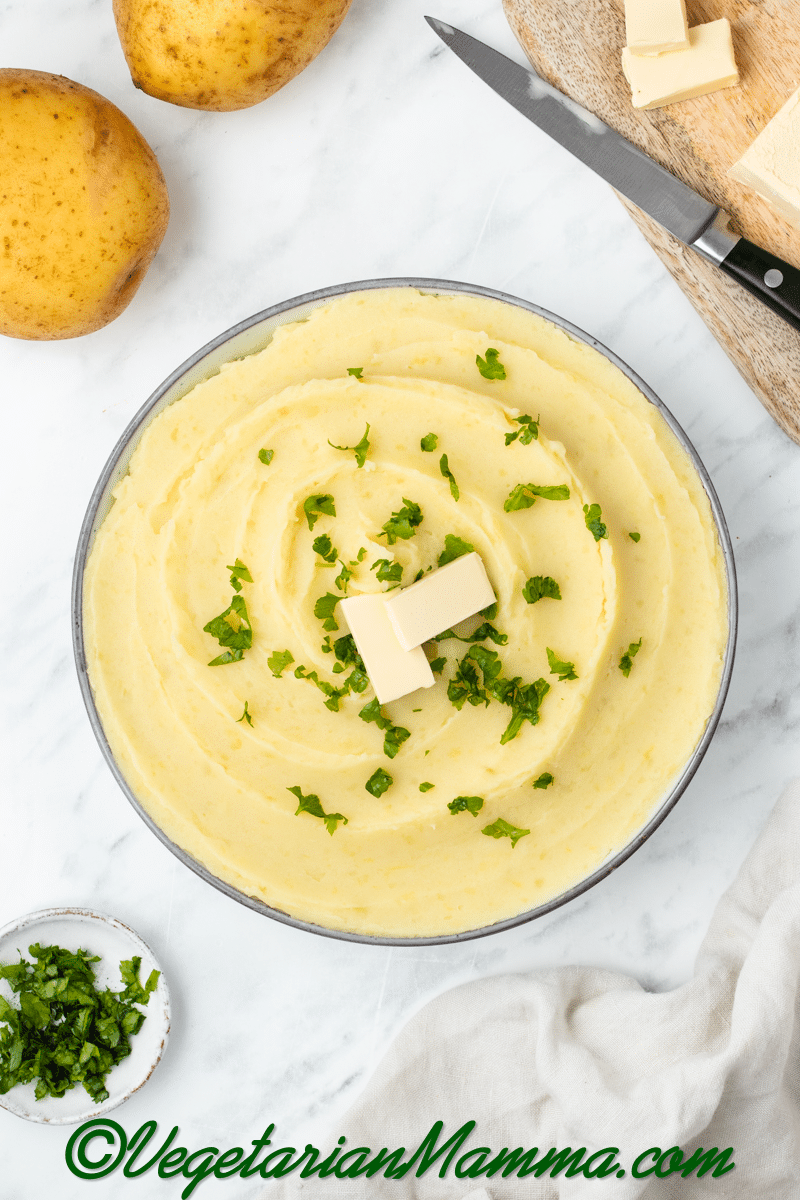 yellow whipped and smooth mashed potatoes with almond milk in a white bowl with two butter rectangles and green herbs on top, knife on brown cutting board to top right, brown washed slightly wet potato to top left, and green herbs to bottom left
