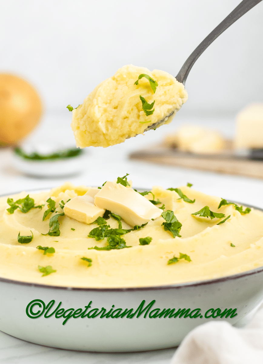spoon full of mashed potatoes and yellow whipped and smooth mashed potatoes with almond milk in a white bowl with two butter rectangles and green herbs on top