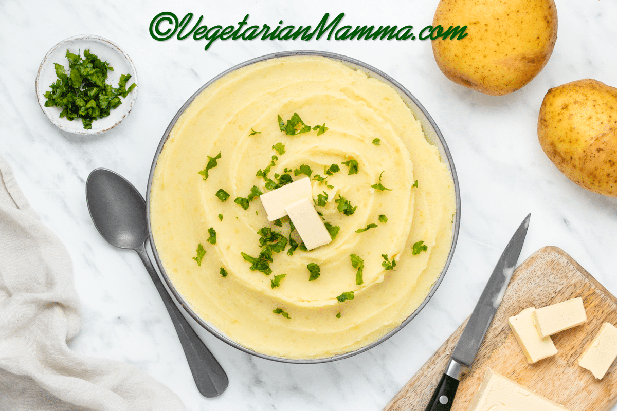 yellow whipped and smooth mashed potatoes with almond milk in a white bowl with two butter rectangles and green herbs on top