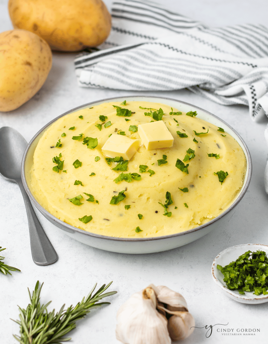 shot of yellow mashed potatoes in a white bowl with two slabs of butter on top and green herbs two brown potatoes to top left, spoon to left, fresh rosemary to bottom left, garlic to bottom right
