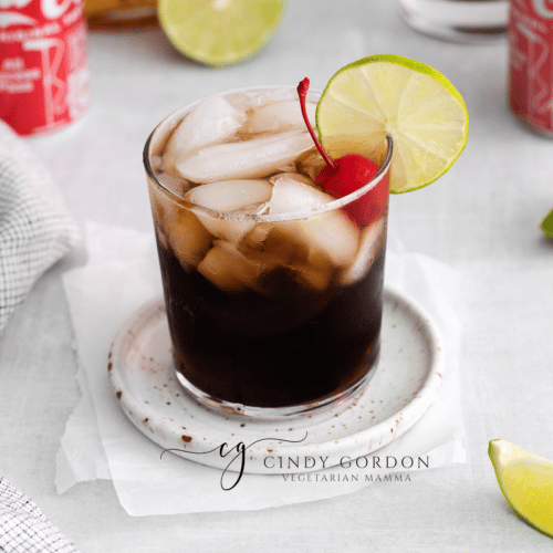 medium clear glass filled with half moon ice cubes and brown liquid. Lime wheel and one cherry in glass