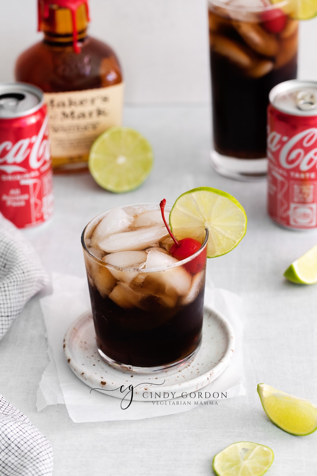 medium clear glass filled with half moon ice cubes and brown liquid. Lime wheel and one cherry in glass coke cans in back along with whiskey bottle Tall glass in background with whiskey in coke