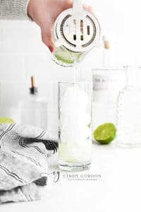 Gin Mojito in a tall clear glass with ice cubes, clear liquid, mint garnish and lime garnish clear liquid being strained into glass