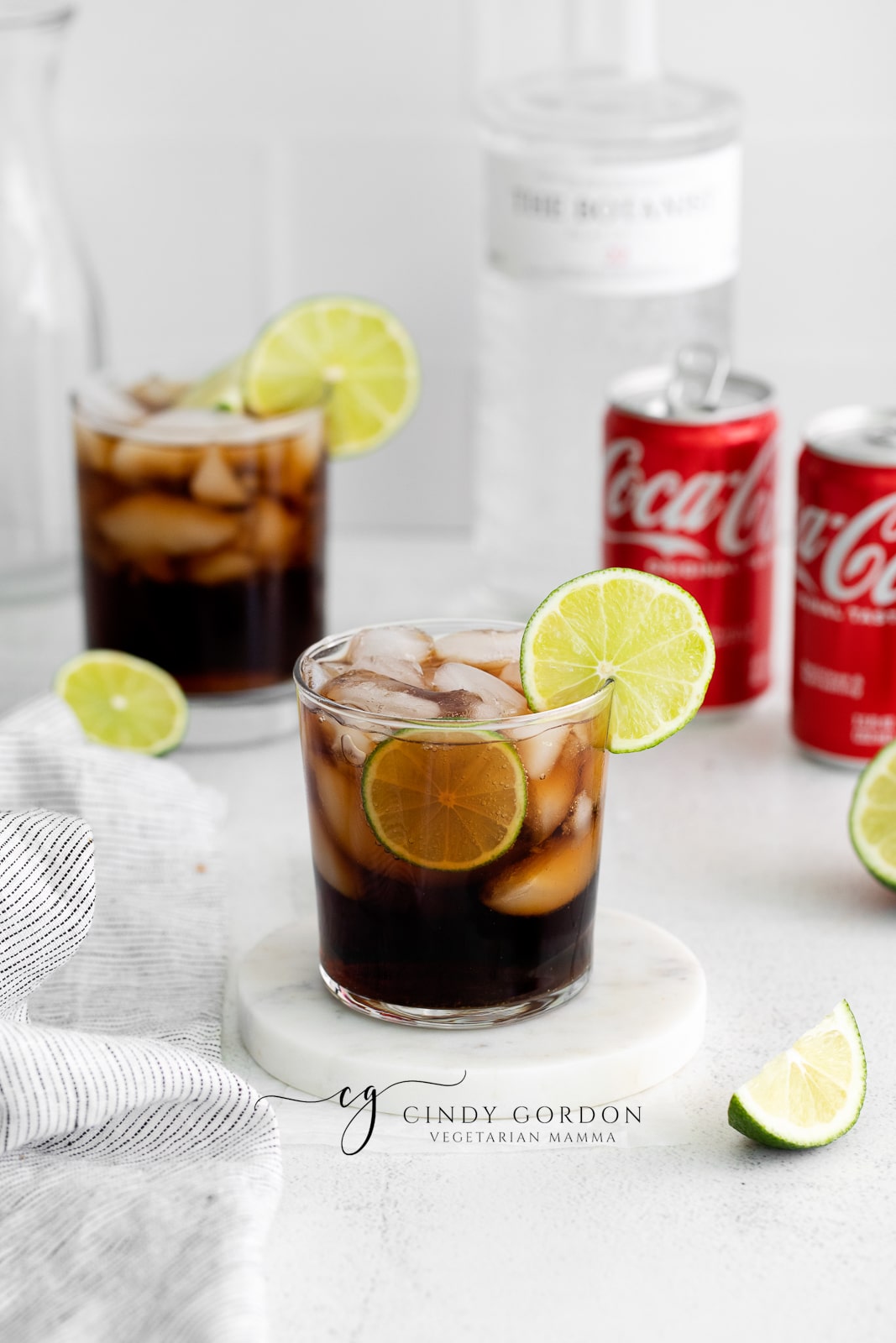 two short clear glasses full of hale moon ice cubes. Brown liquid in glasses with lime wheel on top of glasses. Coke a cola cans in back ground with cut limes and towel to the left. Bottle of gin in the background