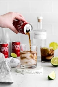 two short clear glasses full of hale moon ice cubes. Brown liquid in glasses with lime wheel on top of glasses. Coke a cola cans in back ground with cut limes and towel to the left. Bottle of gin in the background with a hand pouring brown liquid into glass