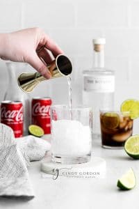 two short clear glasses full of hale moon ice cubes. clear liquid in glasses with lime wheel on top of glasses. Coke a cola cans in back ground with cut limes and towel to the left. Bottle of gin in the background