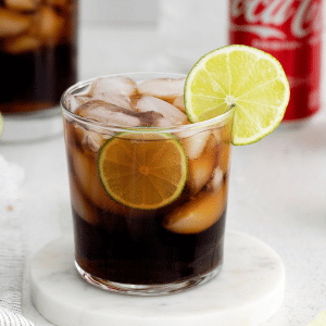 two short clear glasses full of hale moon ice cubes. Brown liquid in glasses with lime wheel on top of glasses. Coke a cola cans in back ground with cut limes and towel to the left. Bottle of gin in the background