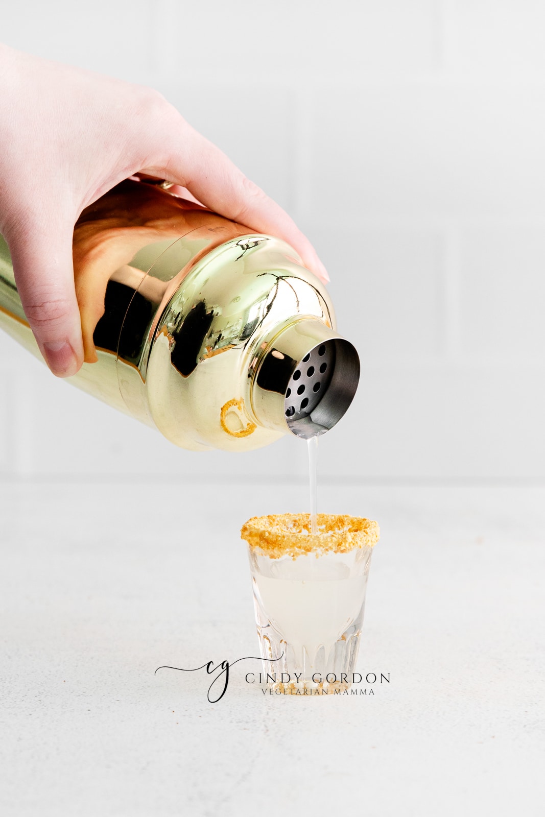 gold cocktail shaker pouring clear liquid into shot glass with brown crumbles on rim