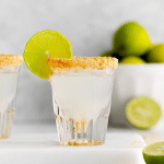 clear shot glass with a milky white clear liquid. Brown crumbled graphams on rim of shot glass. Lime wheel on top side of glass. Limes in backaground