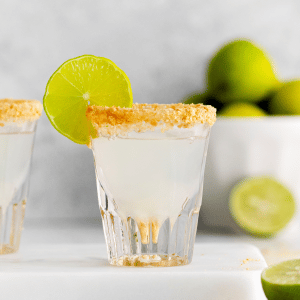 clear shot glass with a milky white clear liquid. Brown crumbled graphams on rim of shot glass. Lime wheel on top side of glass. Limes in backaground