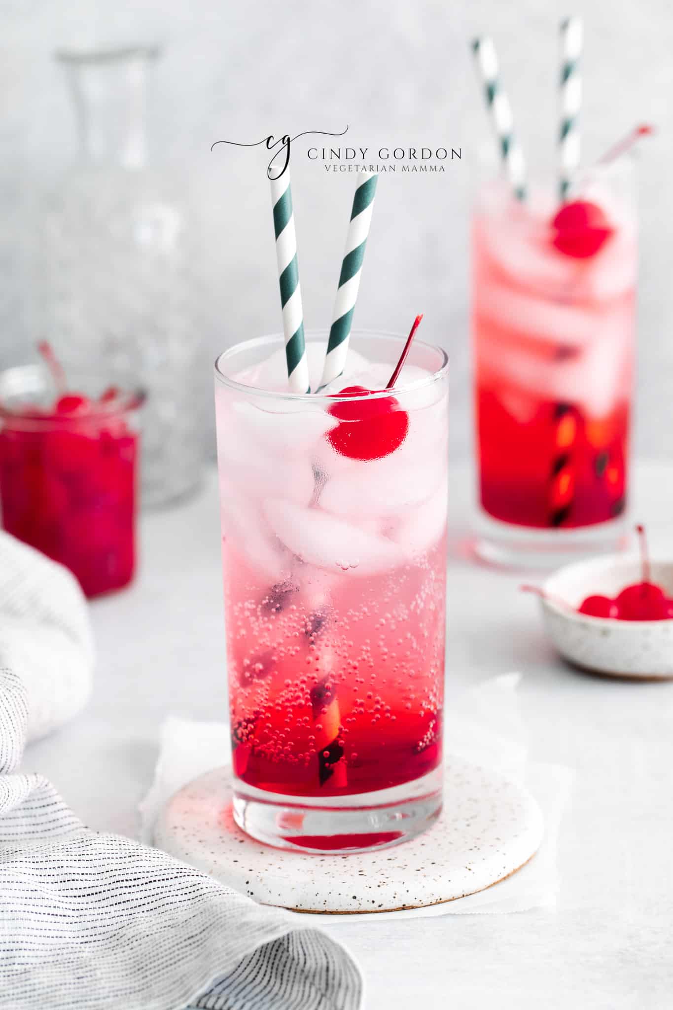 two tall clear glasses filled with red liquid at bottom, clear fizzy liquid at top with ice cubes and two striped straws in each glass. Each glass also has a cherry in it.