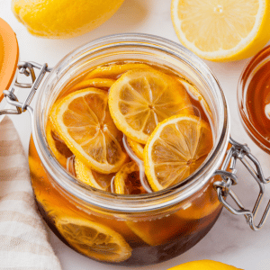 clear jar with lid. Inside are sliced lemons in brown dark honey. slices halved lemons on side and above and honey jar also visable