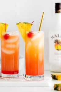 two clear glasses full of malibu barbie drink. Red liquid at bottom, then orange liquid, then half moon ice cubes, a cherry and a piece of pineapple with a bottle of malibu rum in the back right
