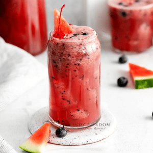 tall clear glass filled with red liquid with black flecks. Tiny watermelon triangle slices, blueberries and cherries. two more glasses in back with same liquid. White napkin to left