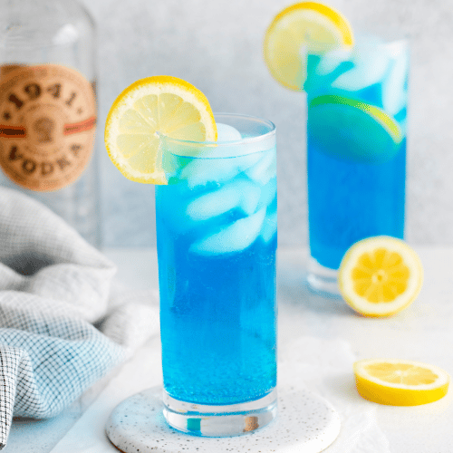 two clear tall glasses filled with ice and a blue liquid, topped with lemon slices, lemon slices also on counter, vodka bottle in back and towel to left side