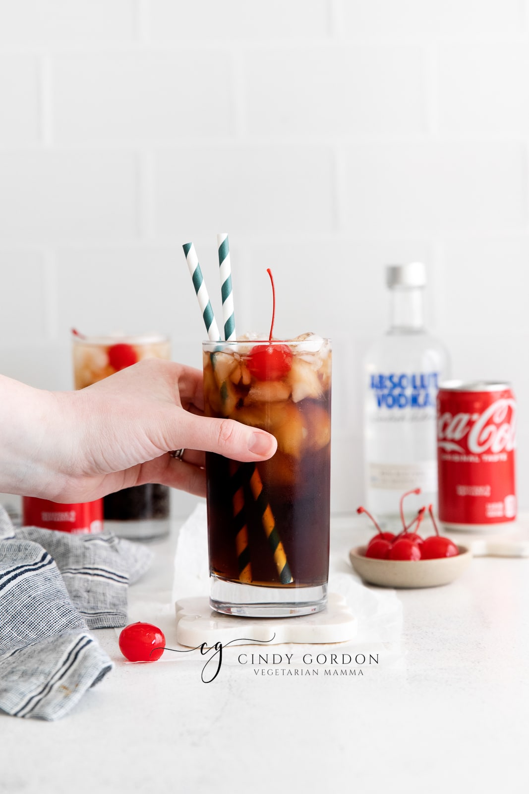hand holding first glass vodka and coke in a tall clear glass with ice and cherries and two straws. Coke can and vodka bottle in back ground. Along with a second vodka coke and a cherry on the table.