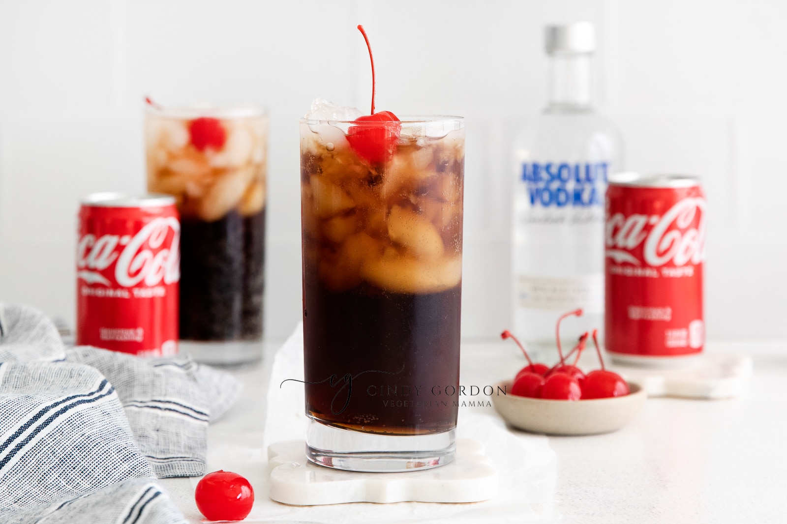horizontal photo vodka and coke in a tall clear glass with ice and cherries and two straws. Coke can and vodka bottle in back ground. Along with a second vodka coke and a cherry on the table.