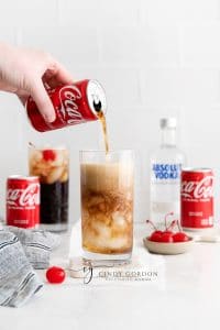 tall clear glass with coke being poured into itand two straws. Coke can and vodka bottle in back ground. Along with a second vodka coke and a cherry on the table.