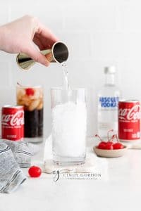 tall glass with clear liquid being poured into it vodka and coke in a tall clear glass with ice and cherr Coke can and vodka bottle in back ground. Along with a second vodka coke and a cherry on the table.