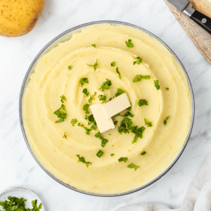 yellow whipped and smooth mashed potatoes with almond milk in a white bowl with two butter rectangles and green herbs on top