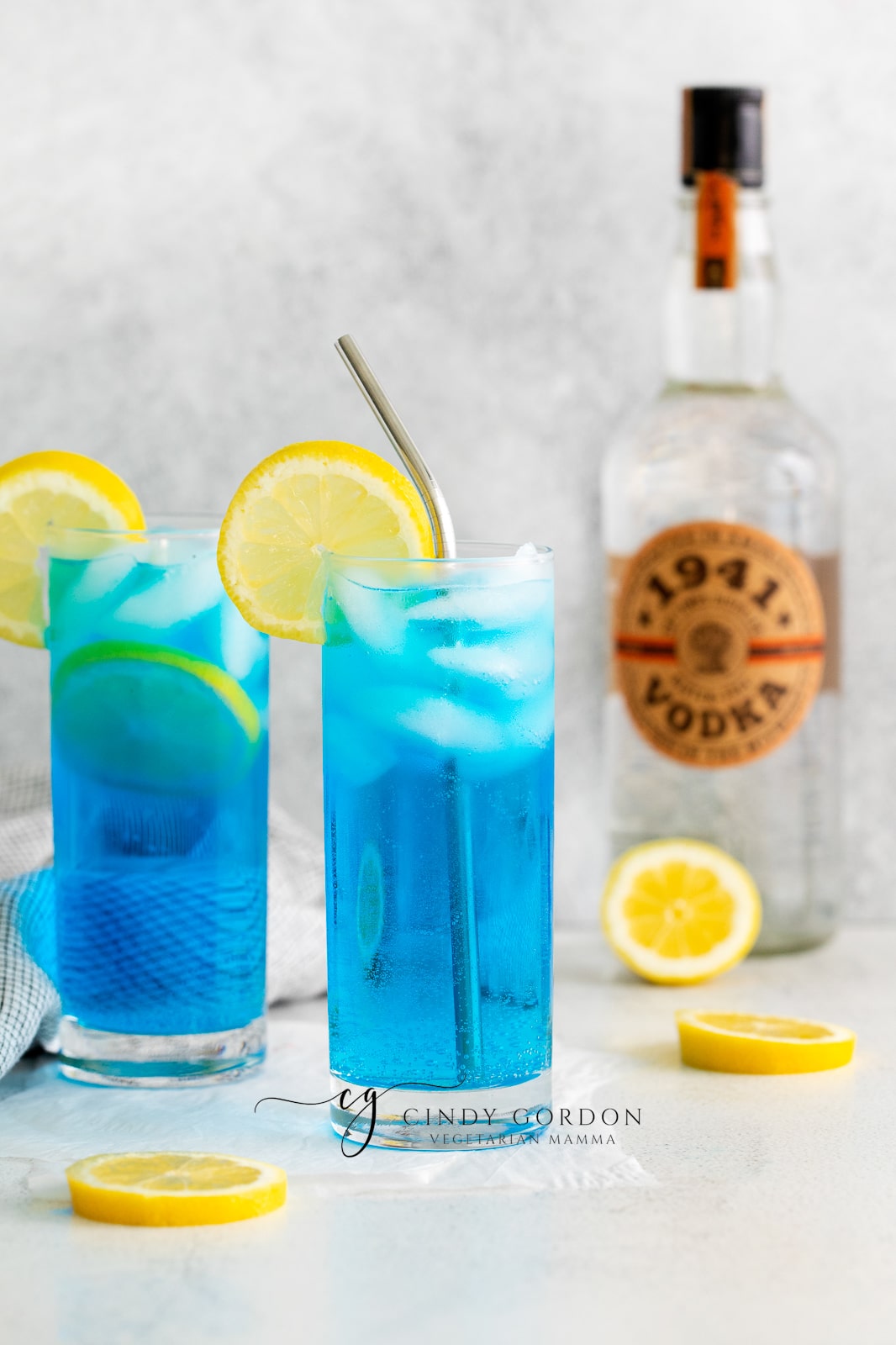 two clear tall glasses filled with ice and a blue liquid, topped with lemon slices, lemon slices also on counter, vodka bottle in back and towel to left side