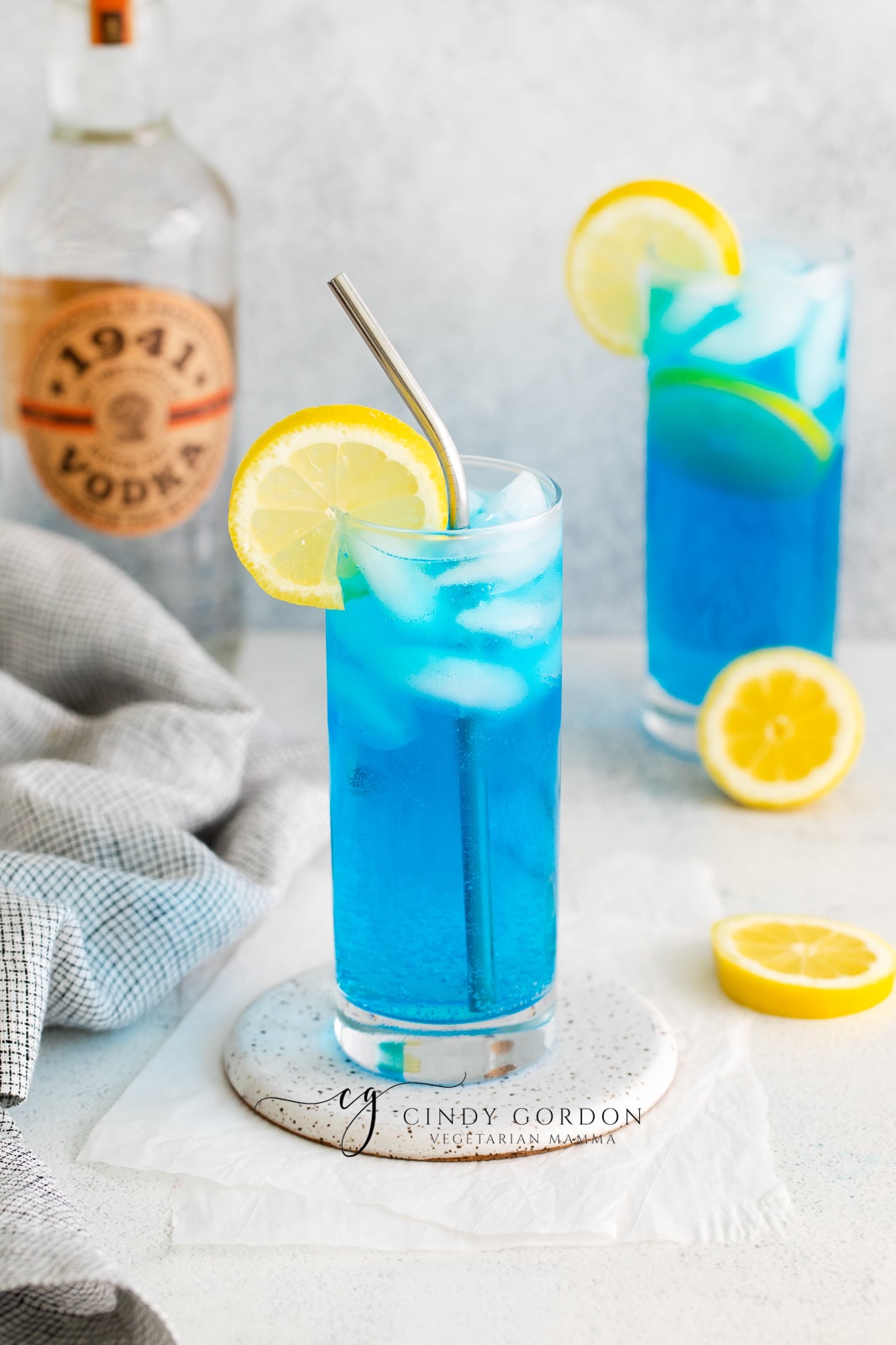 two clear tall glasses filled with ice and a blue liquid, topped with lemon slices, lemon slices also on counter, vodka bottle in back and towel to left side sex on the driveway drink
