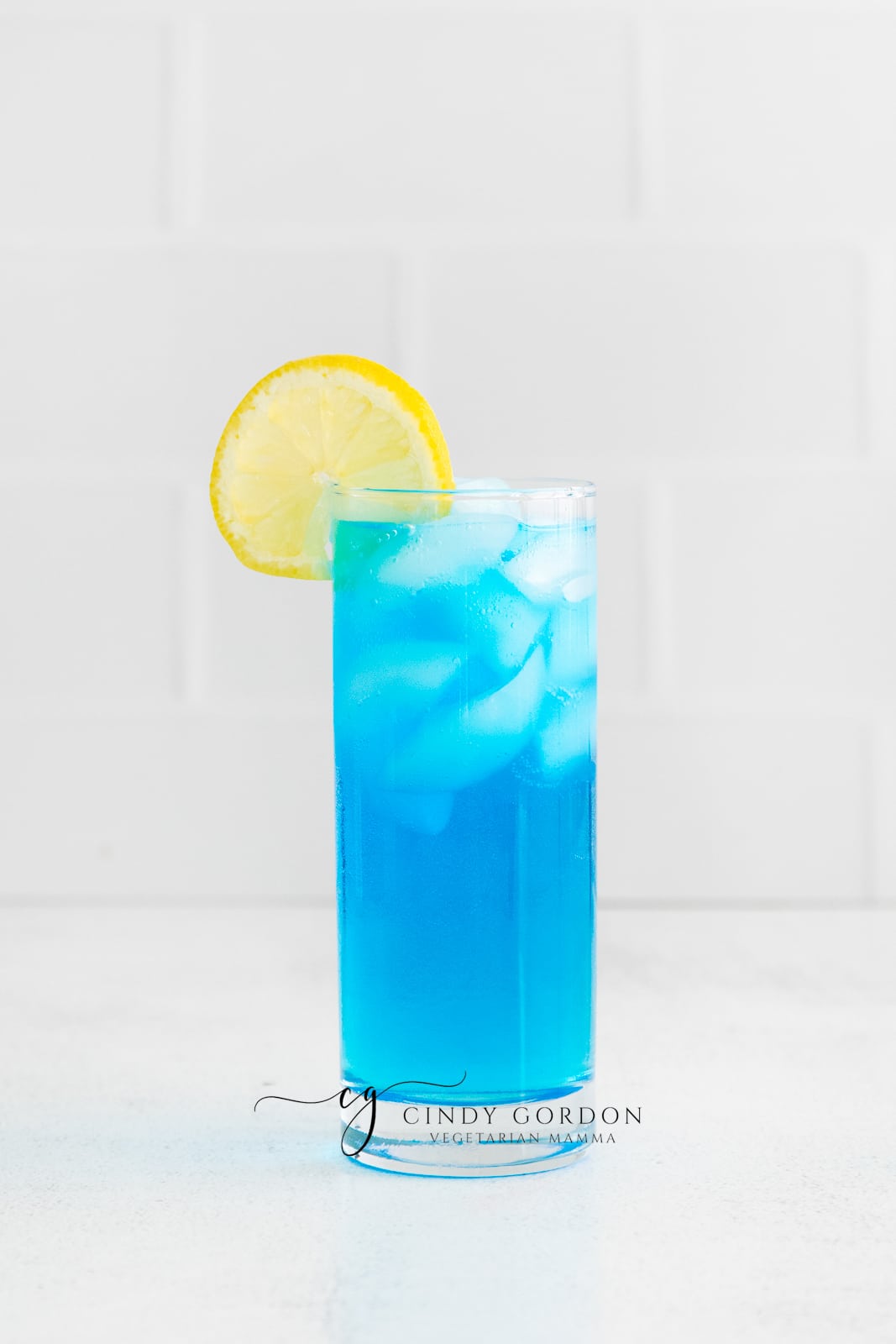 two clear tall glasses filled with ice and a blue liquid, topped with lemon slices