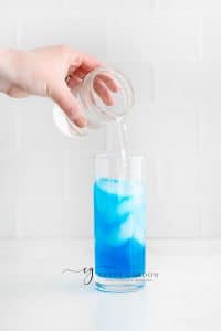 White back ground and white counter, tall clear glass with clear liquid being poured in, there is blue liquid in glass with ice cubes