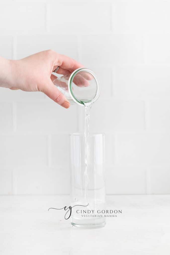 White back ground and white counter, tall clear glass with clear liquid being poured in