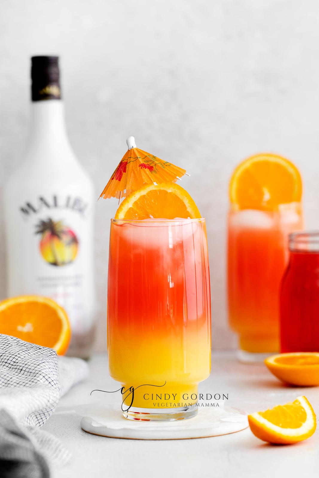 Sex on the beach drink malibu - 2 tall clear glasses filled with half moon ice, orange liquid on the bottom, red liquid on top and orange wheel on very top. Malibu rum bottle in the back. Orange slices on table
