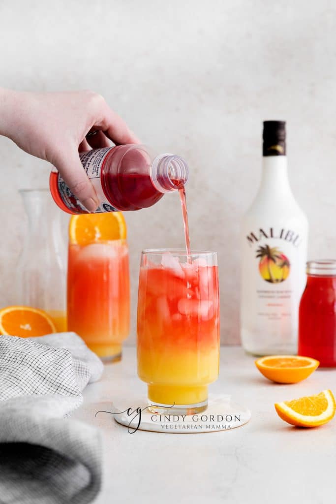 Sex on the beach drink malibu - 2 tall clear glasses filled with half moon ice, orange liquid on the bottom, red liquid on top and orange wheel on very top. Malibu rum bottle in the back. Orange slices on table red liquid being poured in to glass