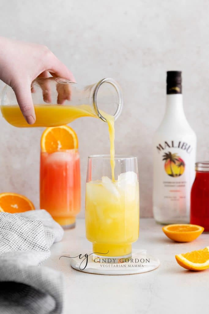 Sex on the beach drink malibu - 2 tall clear glasses filled with half moon ice, orange liquid on the bottom, red liquid on top and orange wheel on very top. Malibu rum bottle in the back. Orange slices on table orange liquid being poured in to glass