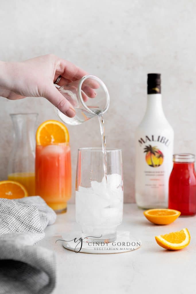 Sex on the beach drink malibu - 2 tall clear glasses filled with half moon ice, orange liquid on the bottom, red liquid on top and orange wheel on very top. Malibu rum bottle in the back. Orange slices on table clear liquid being poured in to glass