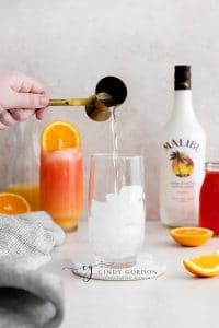 Sex on the beach drink malibu - 2 tall clear glasses filled with half moon ice, orange liquid on the bottom, red liquid on top and orange wheel on very top. Malibu rum bottle in the back. Orange slices on table clear liquid being poured in to glass