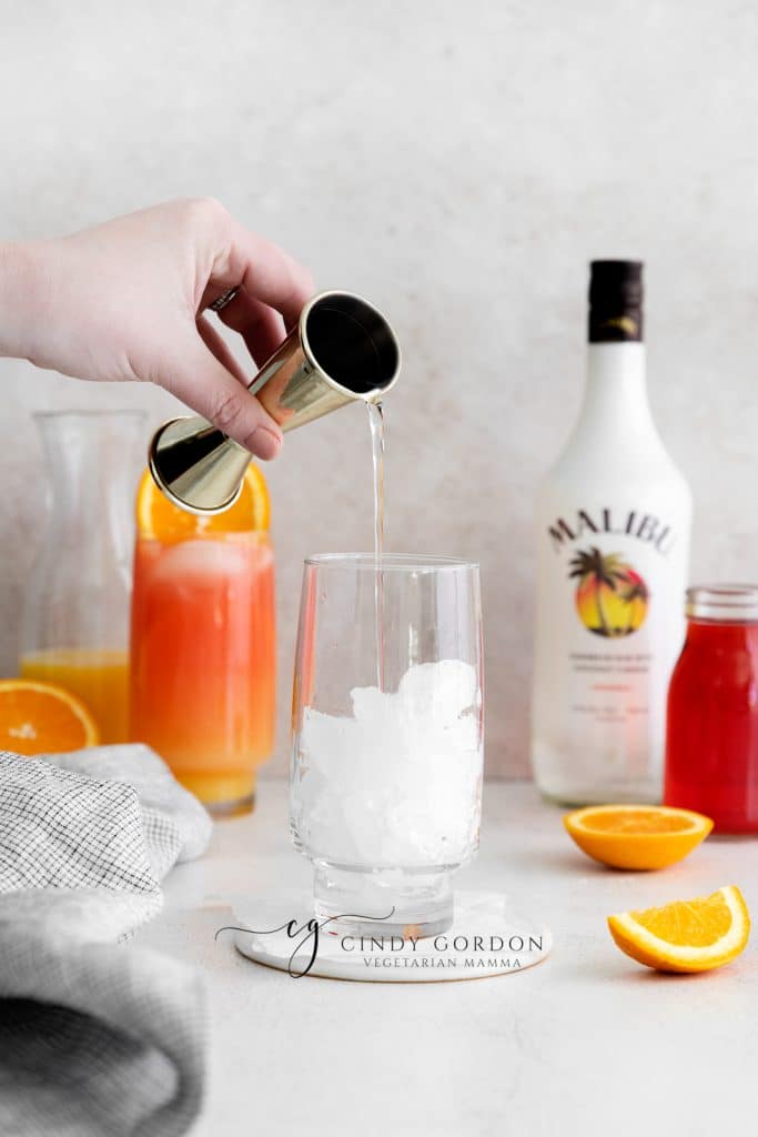 Sex on the beach drink malibu - 2 tall clear glasses filled with half moon ice, orange liquid on the bottom, red liquid on top and orange wheel on very top. Malibu rum bottle in the back. Orange slices on table clear liquid being poured in to glass