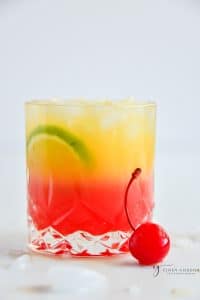 malibu sunrise drink on a rocks glass with red and yellow layers and a maraschino cherry on the side.