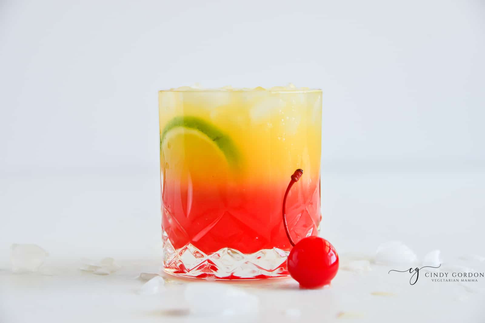 malibu sunrise drink on a rocks glass with red and yellow layers and a maraschino cherry on the side.