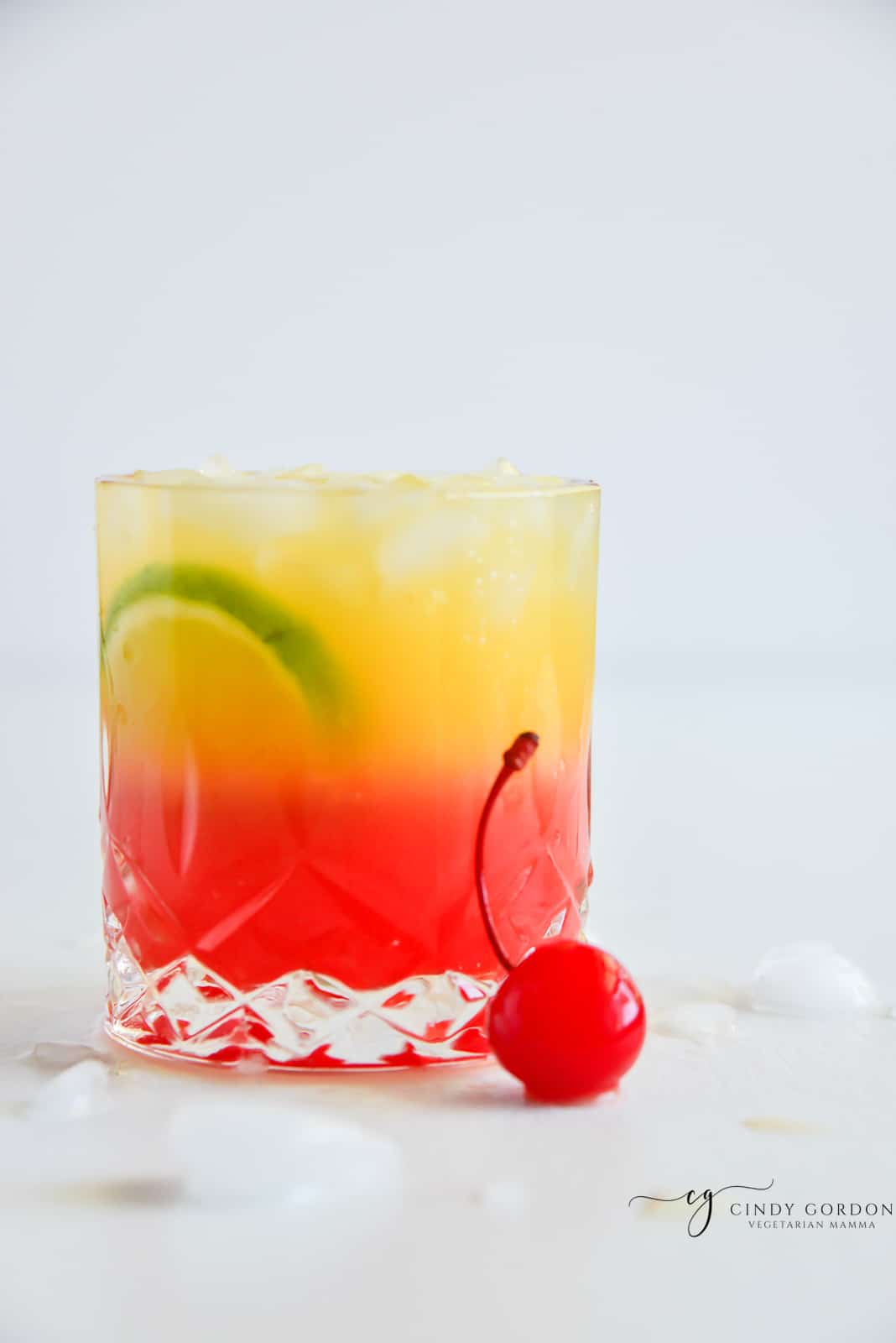 a highball glass with a layered red and yellow malibu sunset drink. Garnished with a cherry and lime slices.
